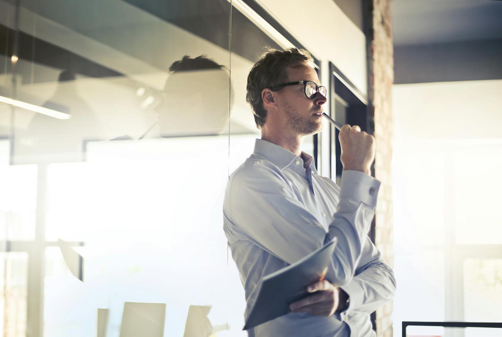 Thoughtful businessman in glasses pondering by office window, holding a folder.